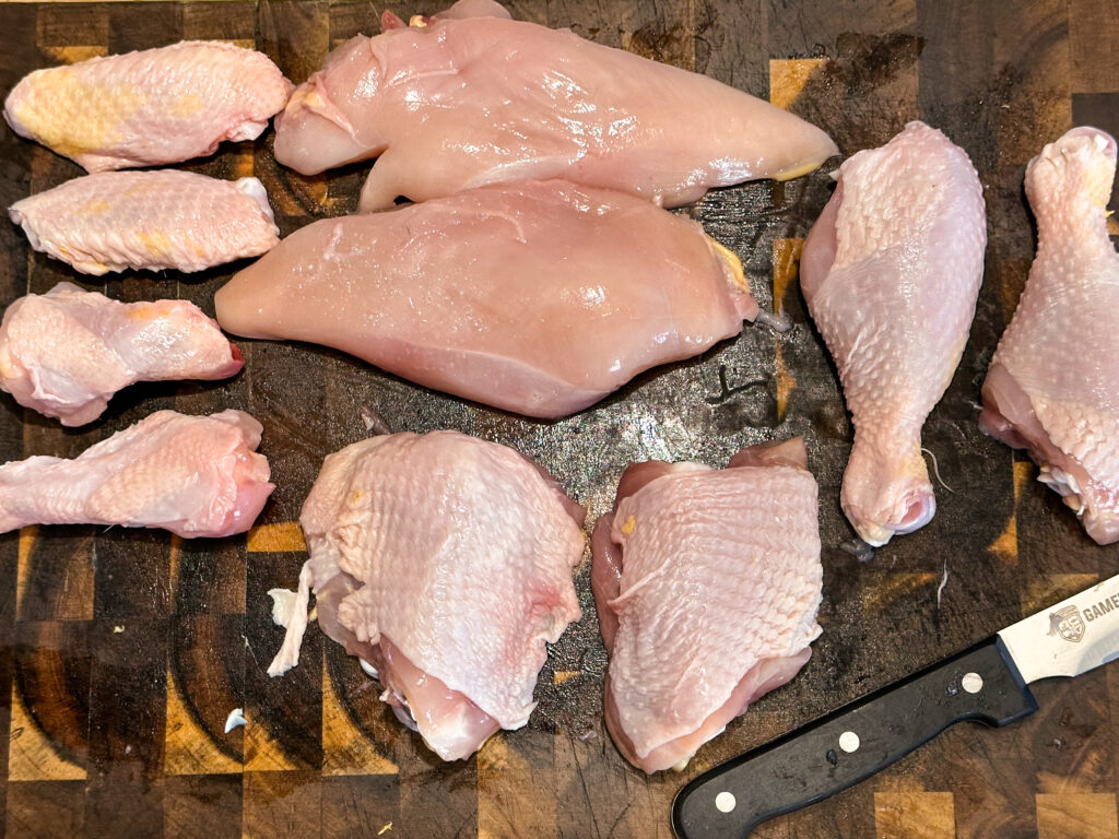 A cutting board with all of the cuts of meat from a whole chicken separated and laid out. Shown are two drumsticks, two thighs, two drumetts, two flats, and two breasts from a whole chicken.