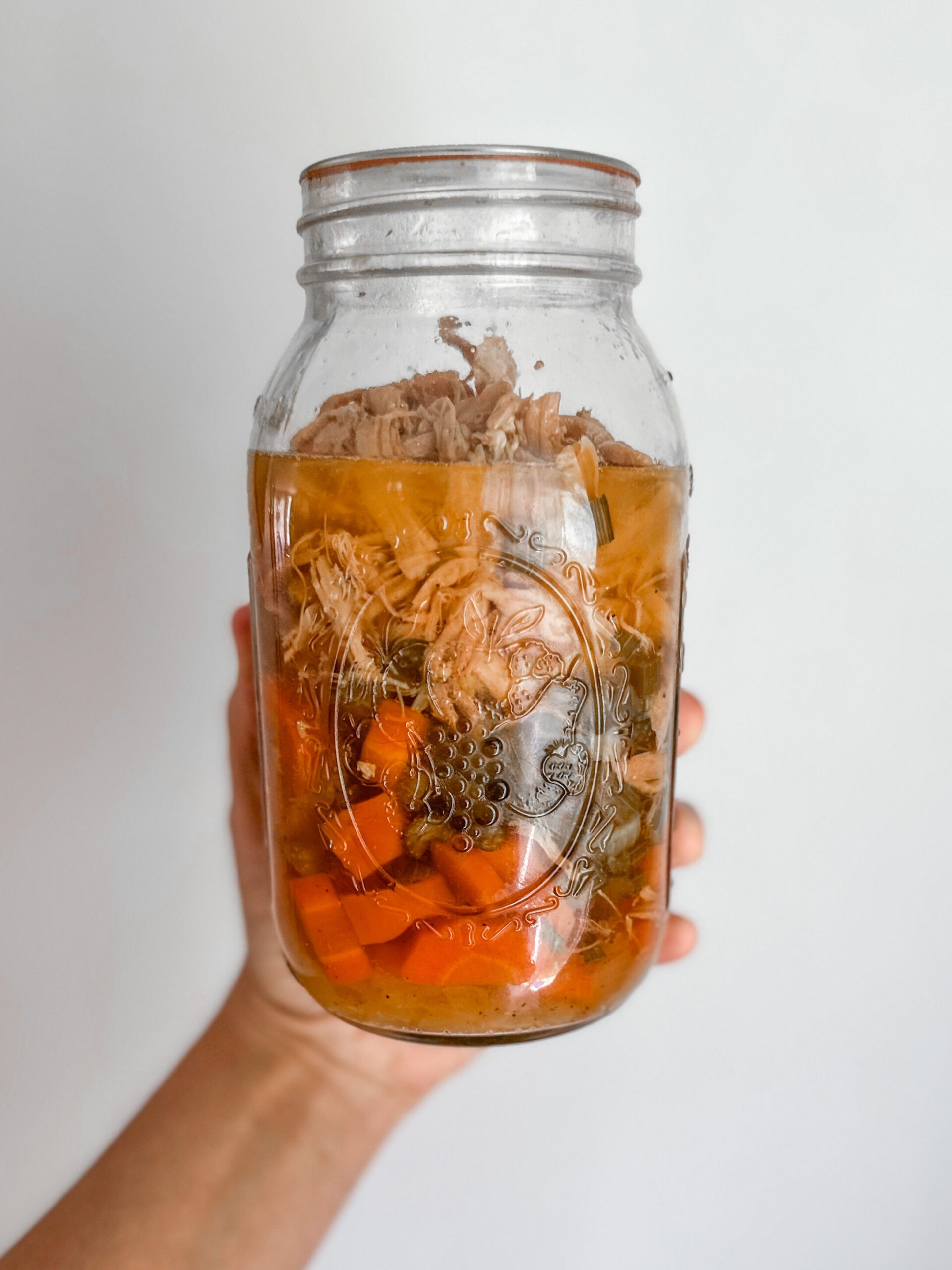 A hand holding a quart sized ball mason jar of homemade, pressure canned chicken soup with carrots and celery. 