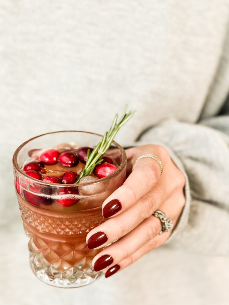 A woman's hand with red holiday nails holding a crystal glass of jingle juice with ice and a cranberry and rosemary garnish. 