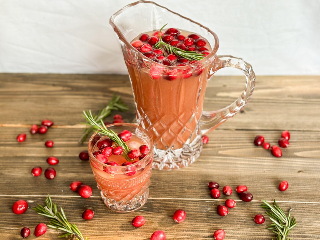 A pitcher and glass of pink holiday punch surrounded by and garnished with fresh rosemary and fresh cranberries.