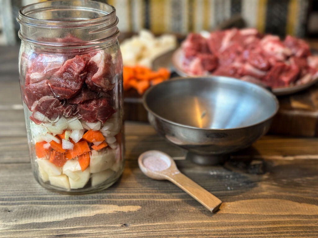 A quart sized ball mason jar filled with raw potatoes, carrots, onions, and chuck roast to make pressure canned beef stew. 