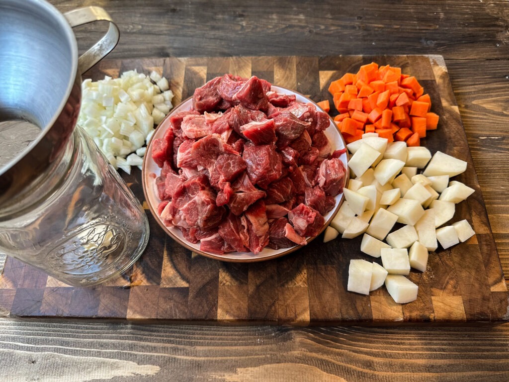 A cutting board with diced onions, carrots, and potatoes. Also shown is a plate of cubed stew beef next to a quart sized canning jar with a canning funnel.
