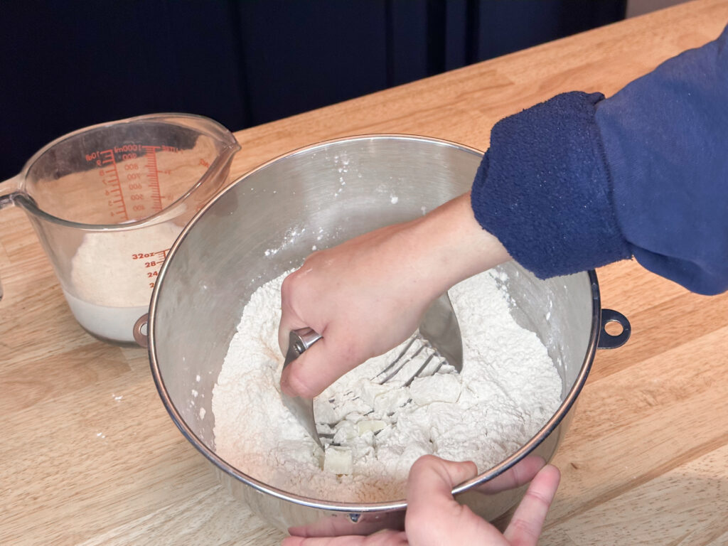 A woman's hands using a pastry cutter to cut butter into a flour mixture in a large, silver mixing bowl.